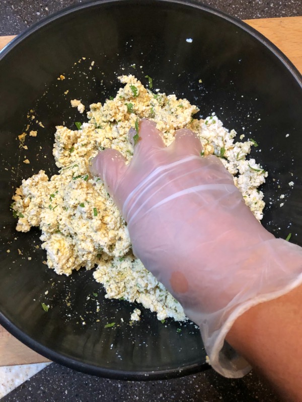 a gloved hand mixing tofu ricotta ingredients in a black bowl.