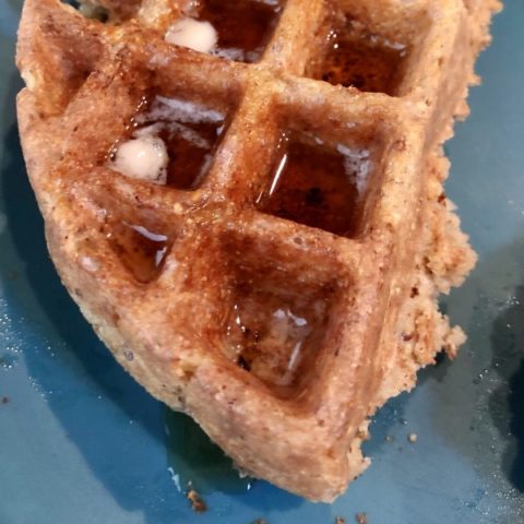 part of a vegan belgian waffle on a blue plate with vegan butter and maple syrup on top.